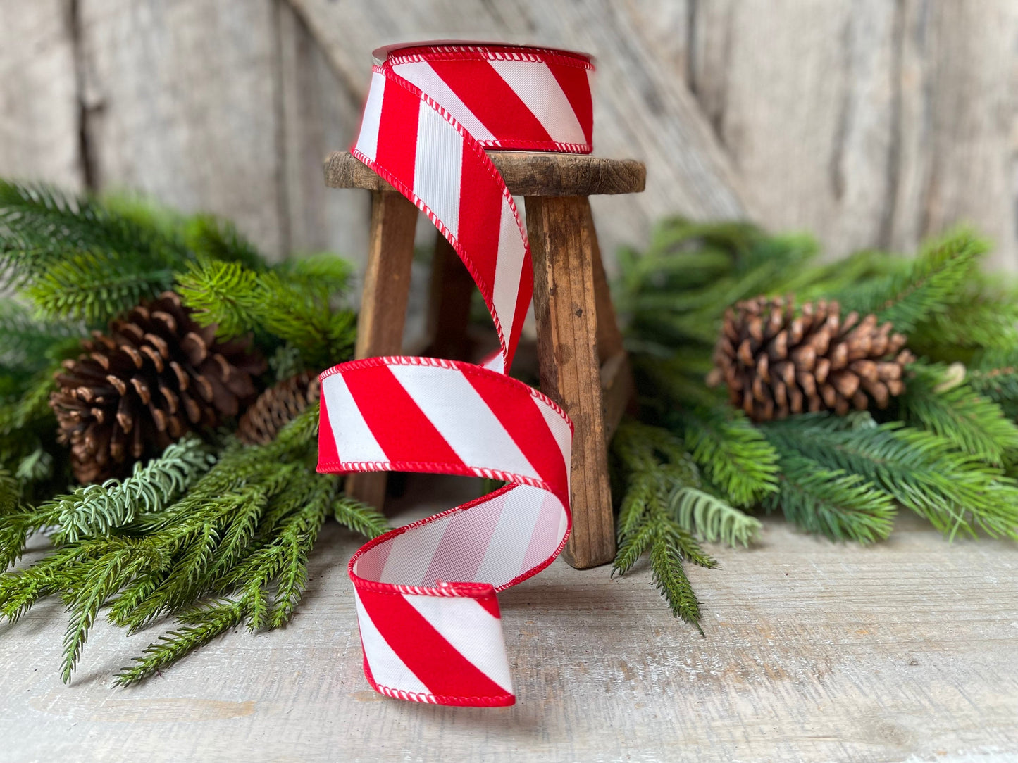 May include: Red and white striped wired ribbon with a candy cane design. The ribbon is displayed on a small wooden stool, surrounded by pine branches and pine cones.  The ribbon is 2.5 inches wide and has a stitched edge. This Christmas ribbon is perfect for crafting, gift wrapping, and decorating.