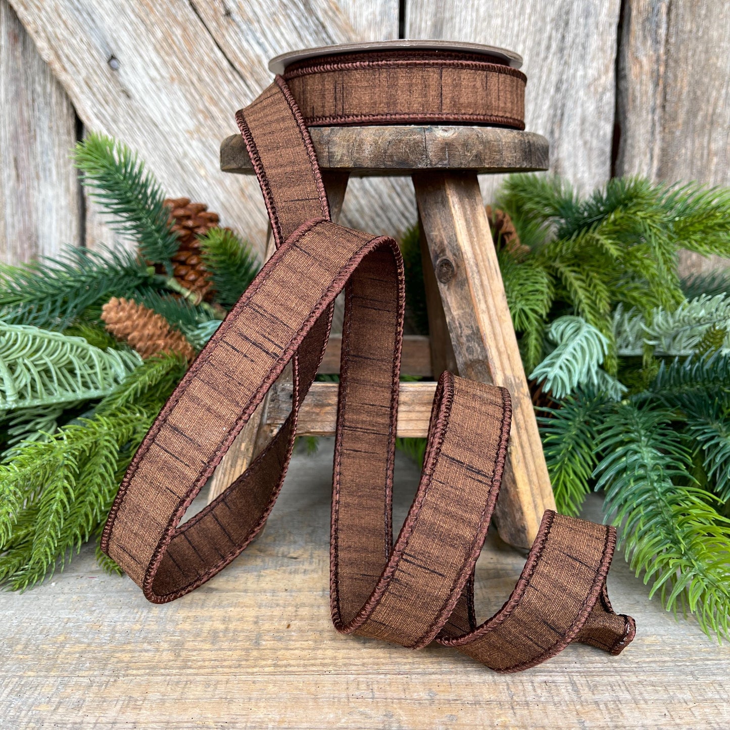 May include: A spool of brown ribbon with a textured pattern and dark brown trim. The ribbon is displayed on a small wooden stool with greenery in the background. The ribbon is unraveled, showing its texture and design, suitable for crafts and decorations.