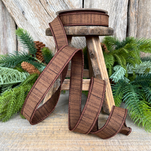 May include: A spool of brown ribbon with a textured pattern and dark brown trim. The ribbon is displayed on a small wooden stool with greenery in the background. The ribbon is unraveled, showing its texture and design, suitable for crafts and decorations.