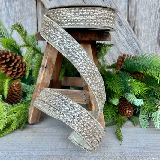May include: A spool of gold and silver ribbon with a beaded design. The ribbon is wrapped around a wooden stool and is surrounded by green foliage.