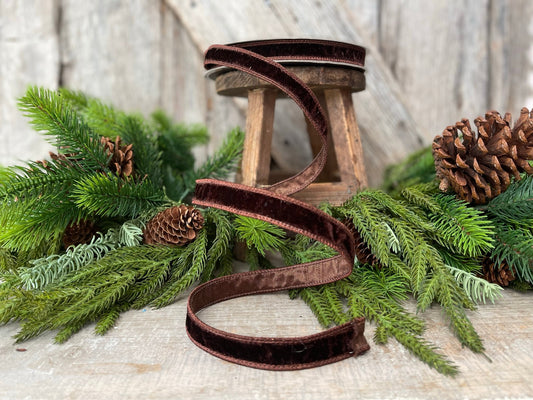 May include: A brown velvet ribbon with a slight sheen. The ribbon is wrapped around a small wooden stool and is laying on a wooden surface with green pine branches and pine cones.