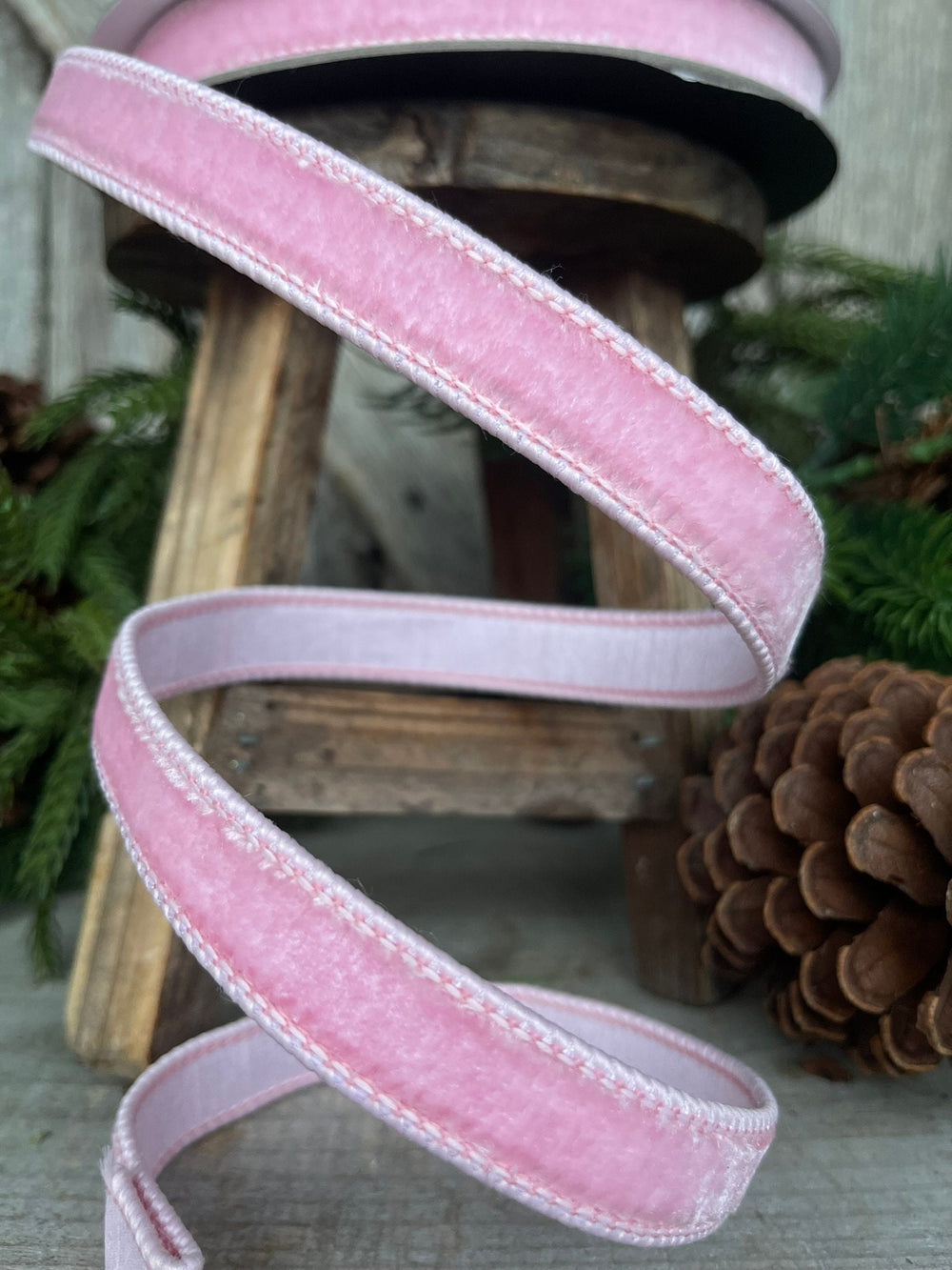 May include: Pink velvet ribbon with a white stitched edge. The ribbon is rolled up and displayed on a wooden stool.
