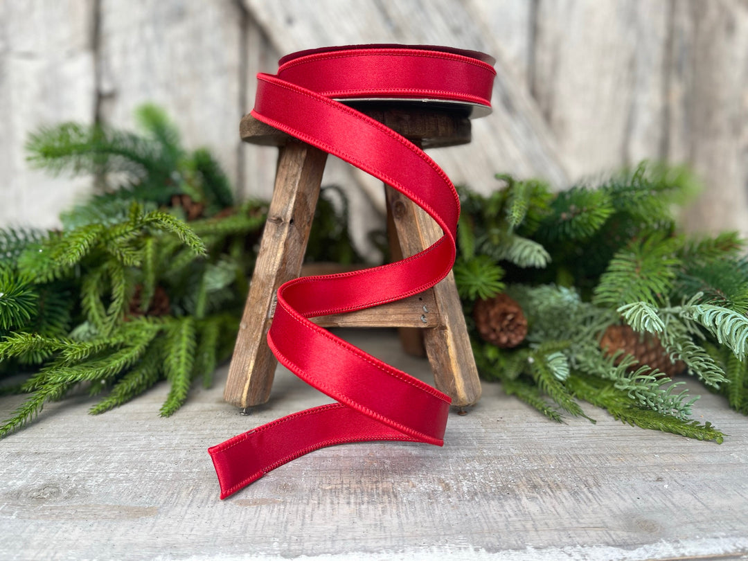 May include: A spool of vibrant red satin ribbon, partially unrolled, sits on a small wooden stool. The ribbon's glossy texture is highlighted against a backdrop of evergreen branches and a rustic wooden surface, suggesting a festive or craft theme.